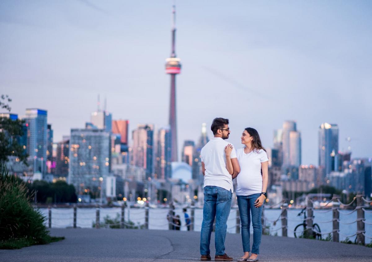 A couple posing on the waterfront with the iconic CN Tower and Toronto skyline in the background for their maternity photo session in Toronto.