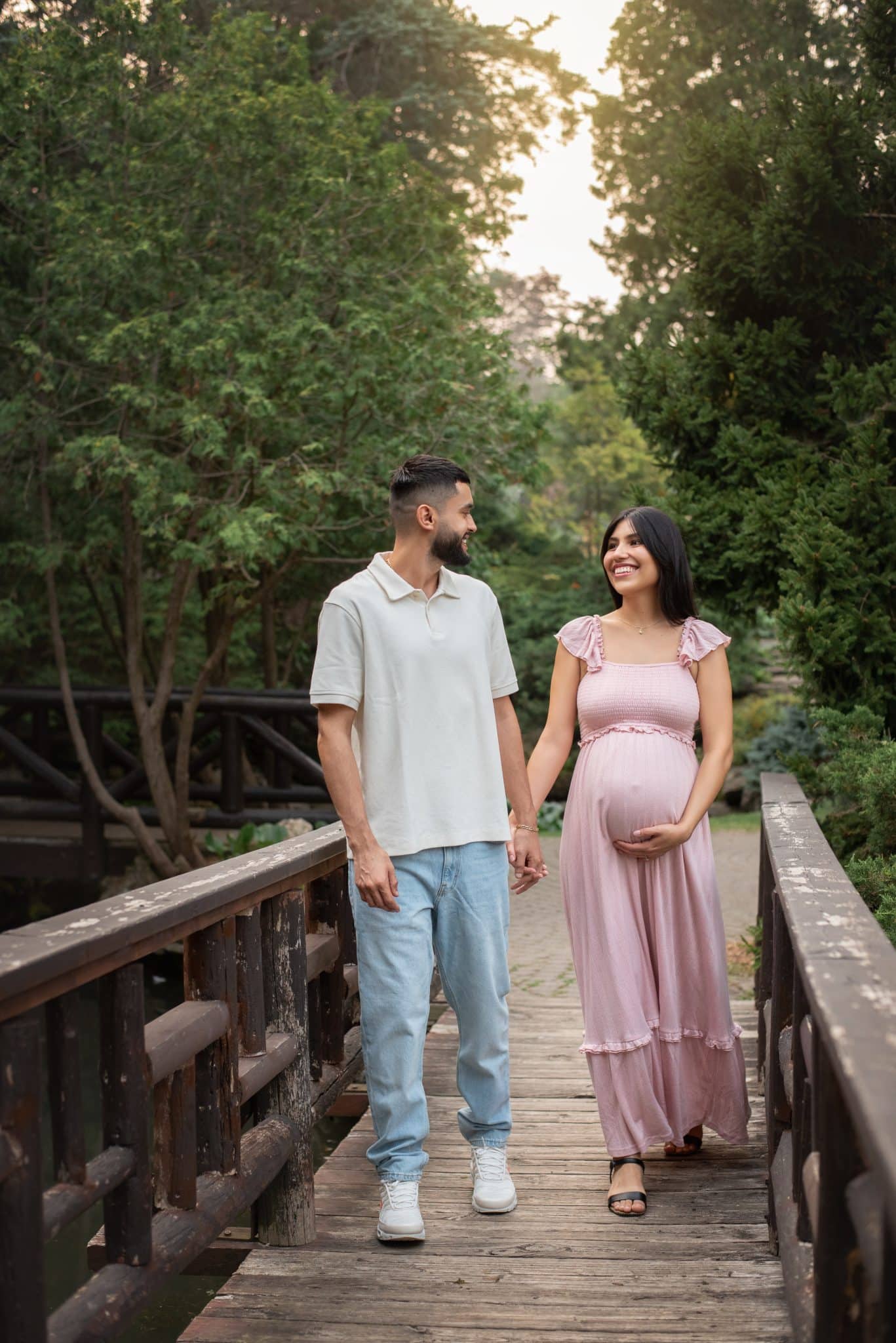 An expectant couple walking hand-in-hand across a rustic wooden bridge during an outdoor maternity photo session in Toronto.