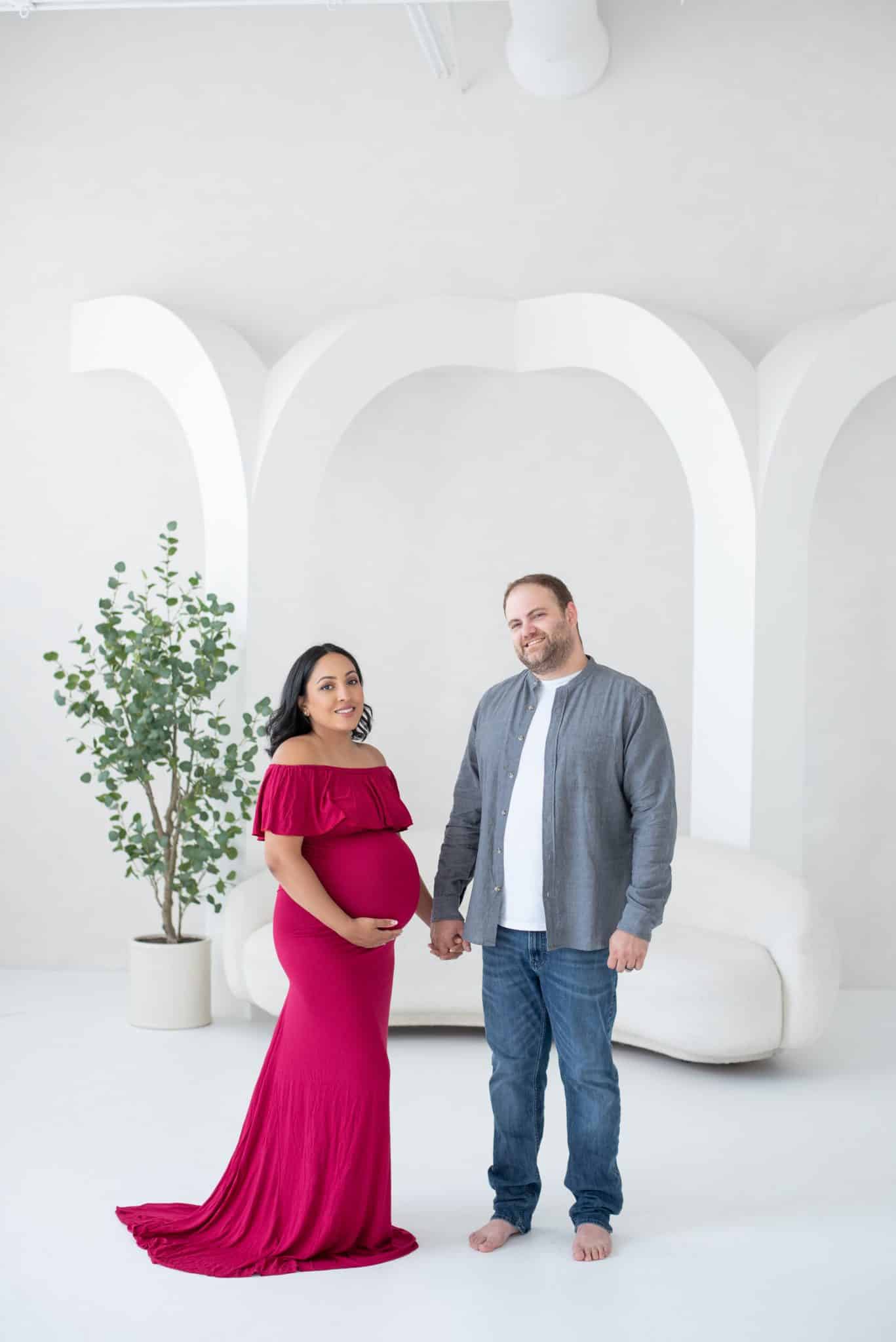A full-length studio portrait of an expectant couple, with the woman in a vibrant red dress, captured during a professional maternity photo session.