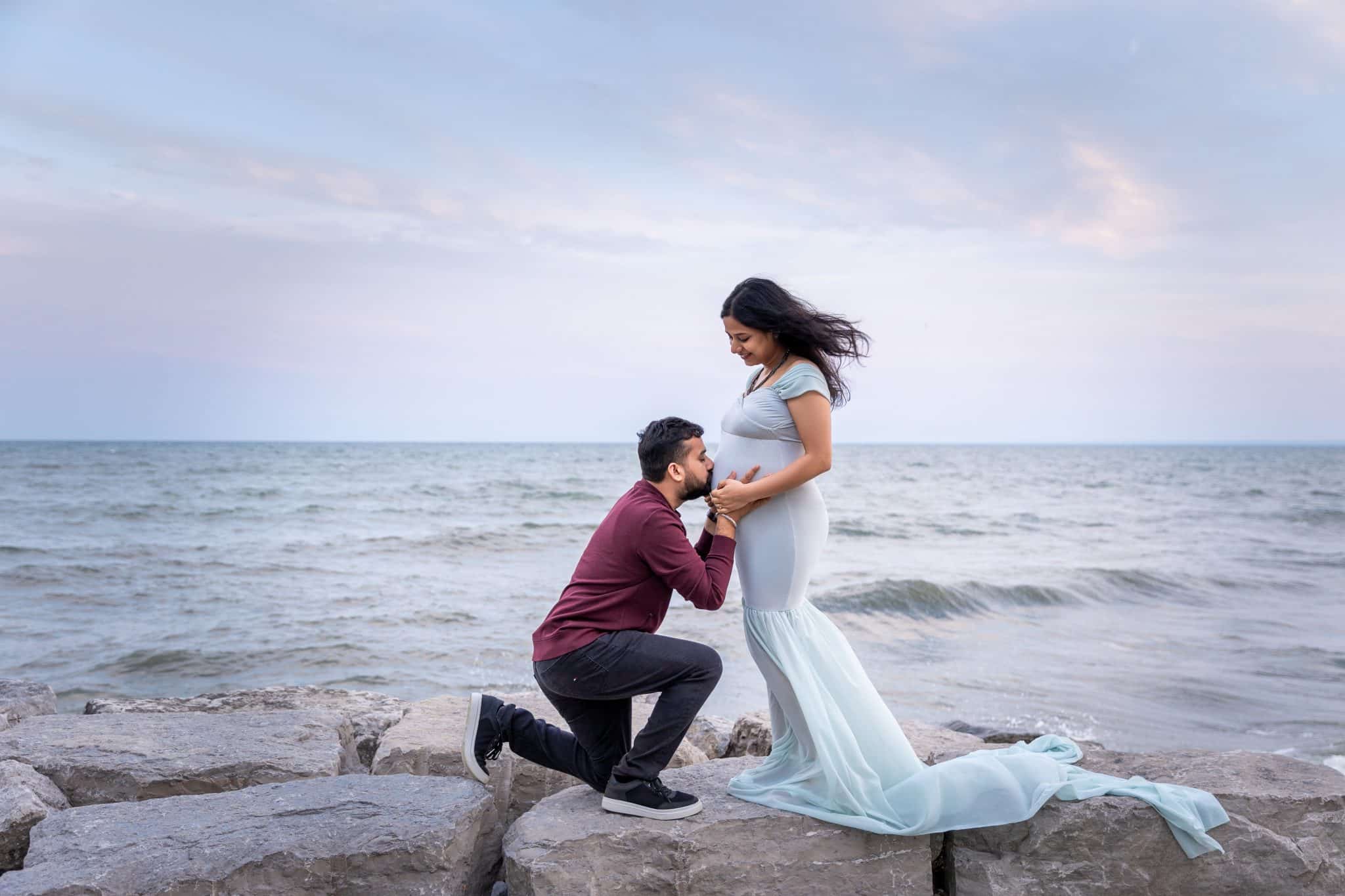 A man kneeling to kiss his pregnant partner's belly on a rocky beach at dusk during a romantic maternity photo session in the GTA.