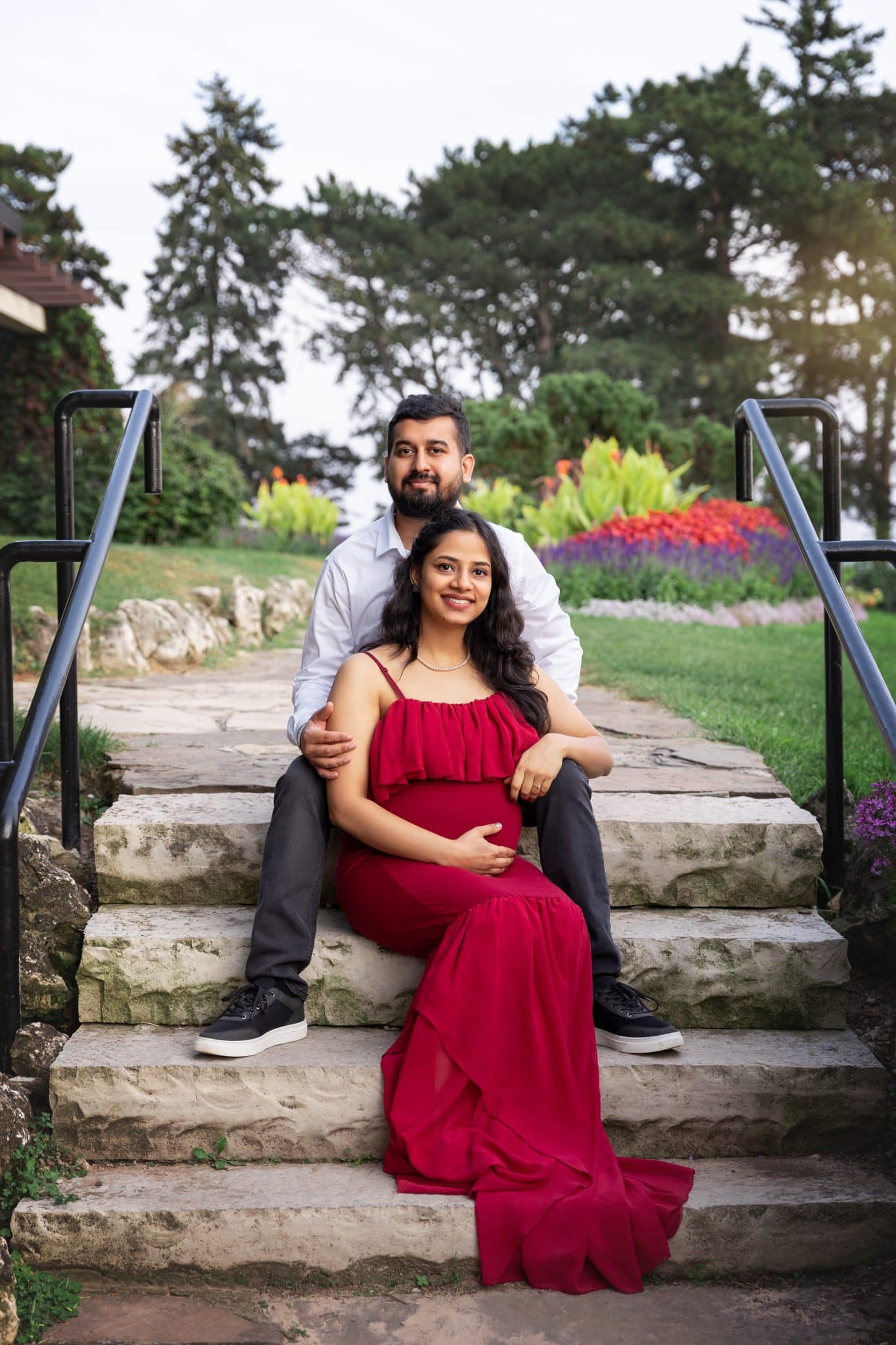 An expectant couple sitting on stone steps in a formal garden, with the woman in a flowing red dress for a pregnancy photoshoot Brampton.