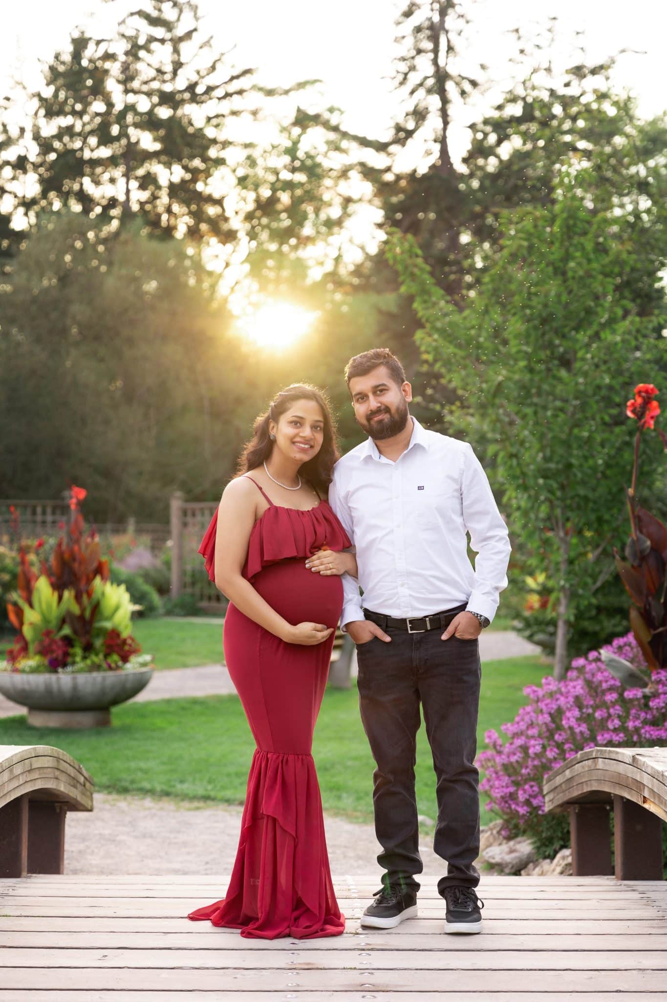 A pregnant woman in a vibrant red gown posing with her partner in a scenic garden at sunset for a maternity photo session Mississauga.