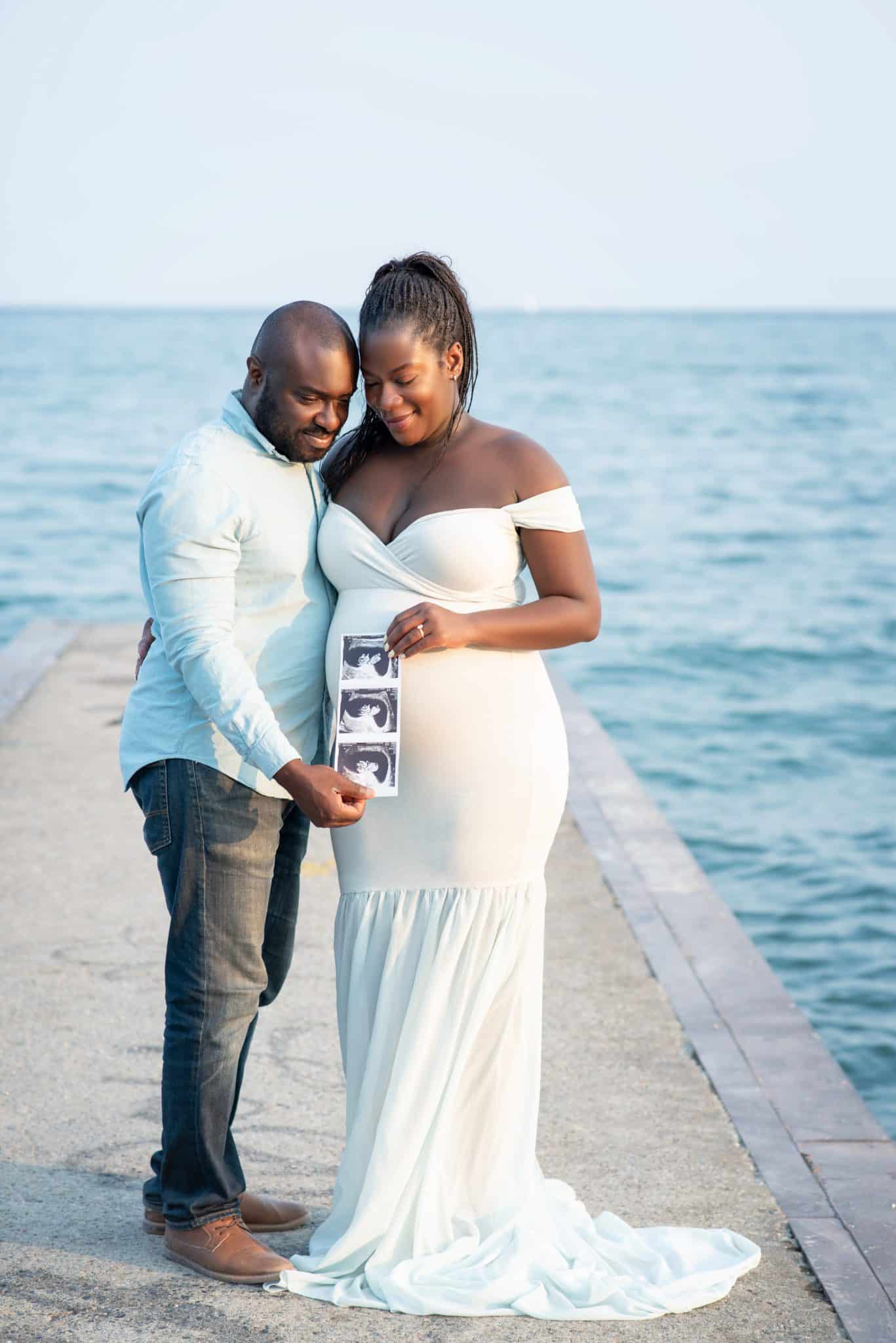A pregnant woman in a white gown and her partner posing on a pier overlooking the water for their pregnancy photoshoot Brampton.