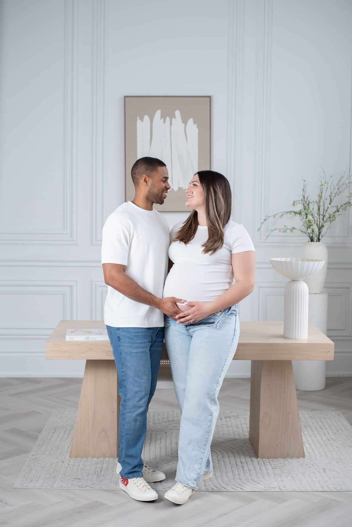 A full-length studio portrait of a pregnant woman and her partner posing in a bright, minimalist setting for a professional maternity photoshoot.