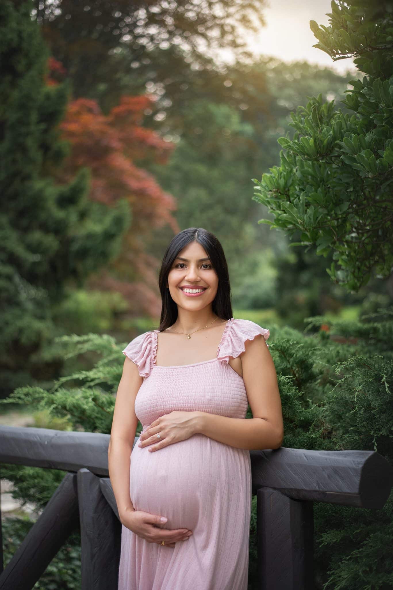 A pregnant woman in a soft pink dress smiling while cradling her belly in a lush, sun-drenched garden during a maternity photo session Mississauga.