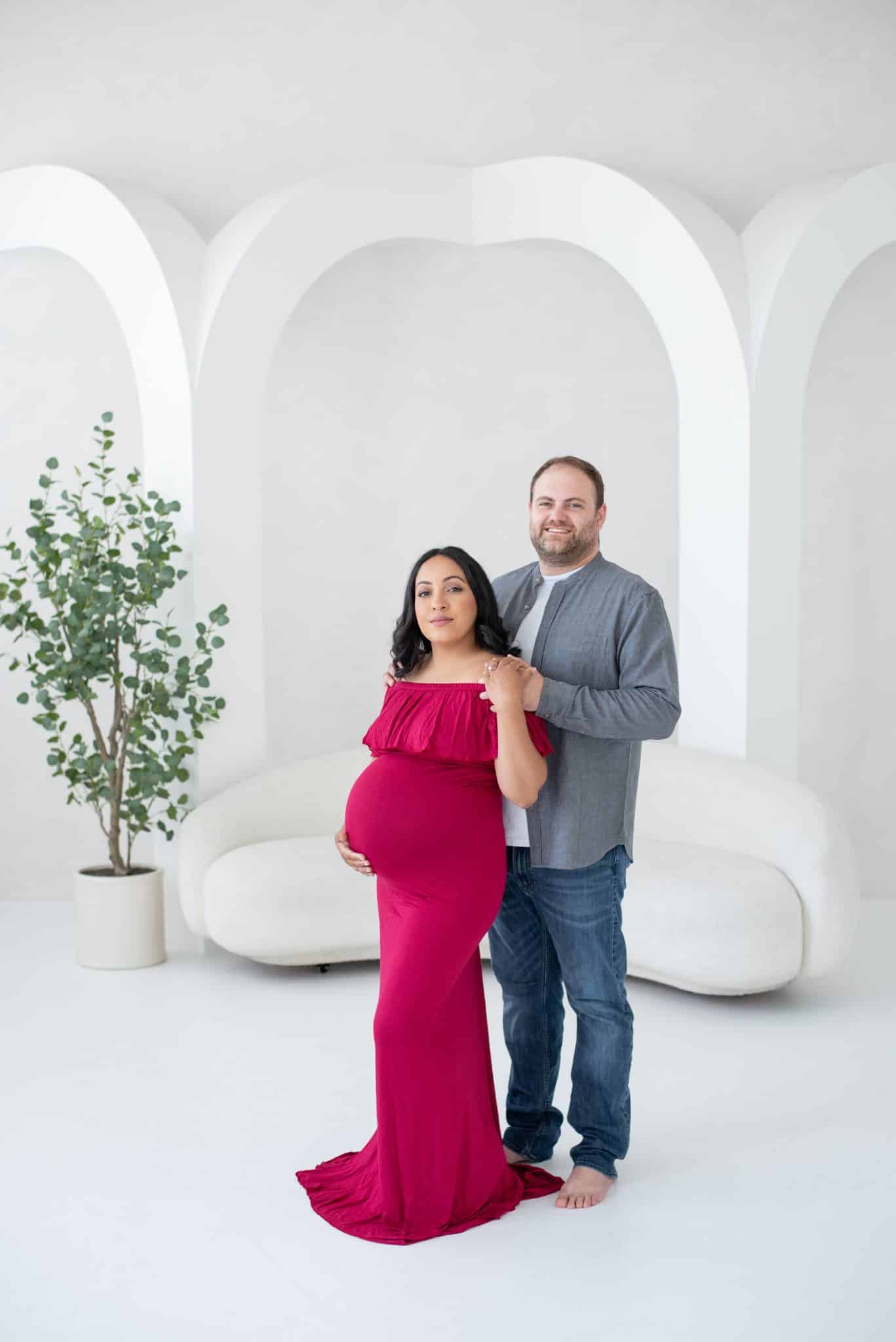 A pregnant woman in a stunning red gown posing with her partner in a minimalist studio for her pregnancy photoshoot Brampton.