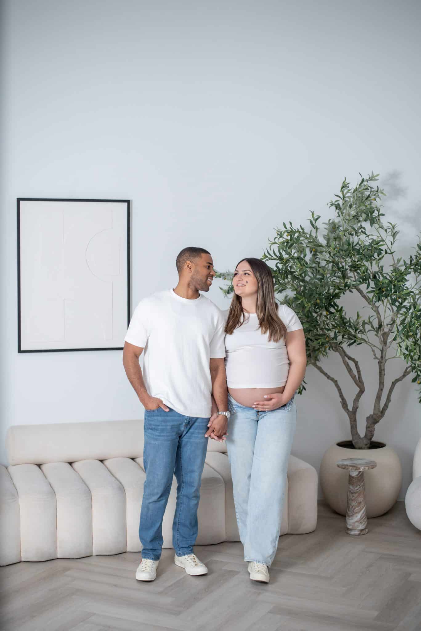 An expectant couple standing together in a clean, modern studio space during their indoor maternity photo session in Toronto.