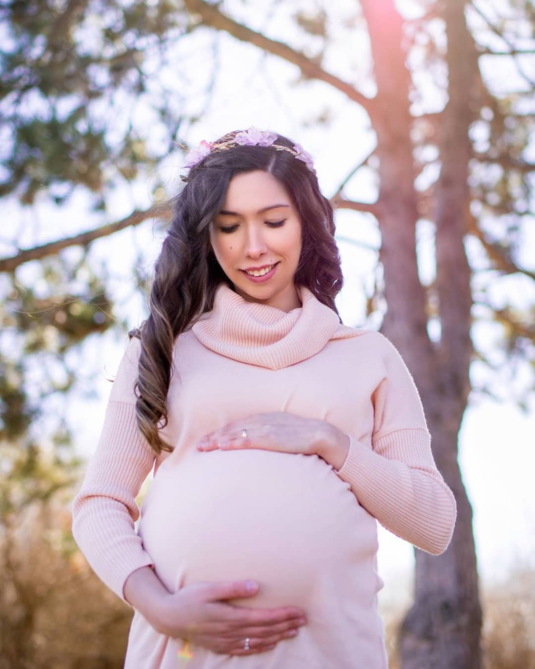 A pregnant woman wearing a soft pink sweater dress posing in a sunlit wooded area for her outdoor pregnancy photoshoot Brampton.